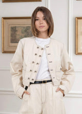 Person wearing a cream-colored jacket with a white shirt and pants, standing against a wall with framed pictures.